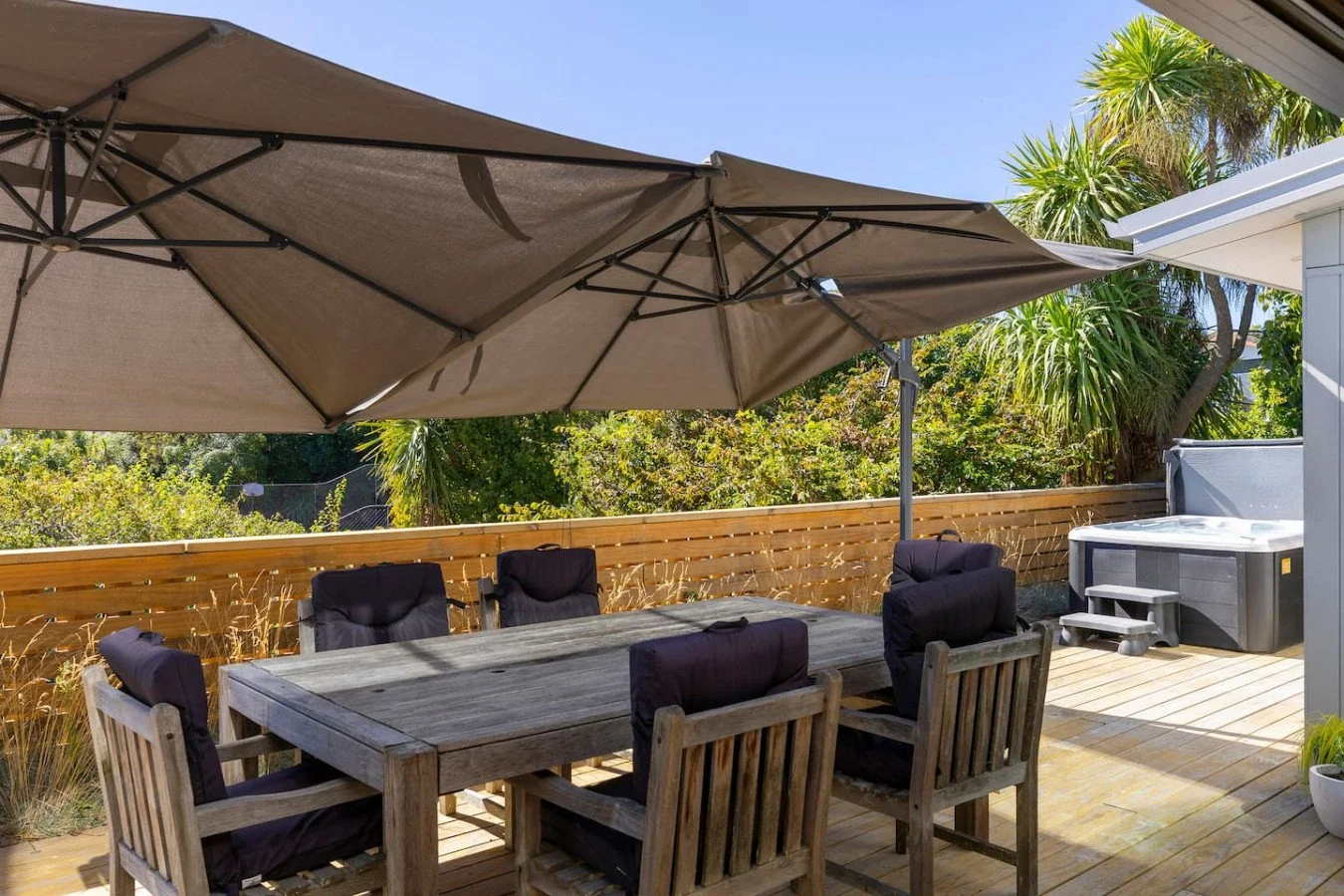 Outdoor wooden patio deck with a rectangular table and six chairs, two with black cushions, shaded by large beige umbrellas, surrounded by a wooden fence, with lush green trees and plants in the background, and a hot tub on the right side.