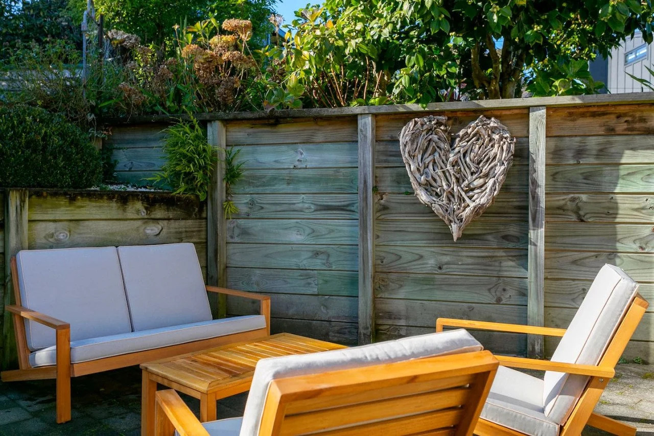 A cozy outdoor patio with wooden and cushioned furniture, a wooden fence decorated with a driftwood heart, lush green plants, and trees in the background.