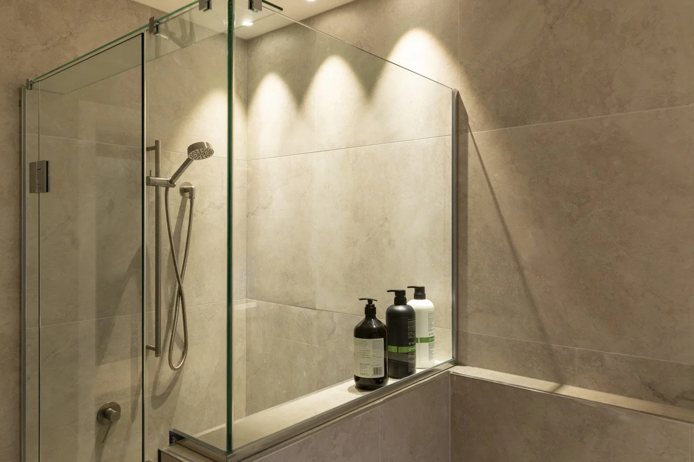 Empty shower area with beige tiled walls, a handheld showerhead, and three bottles of toiletries on a ledge inside the glass enclosure.