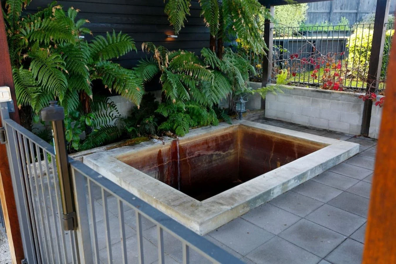 Empty copper hot tub with water draining out, surrounded by potted plants and a metal fence in a backyard.