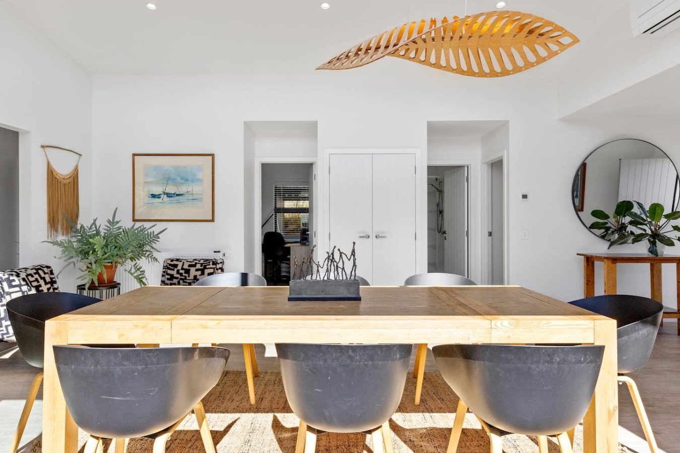 Modern dining room with a wooden table and black chairs, white walls, a round mirror, indoor plants, artwork, and natural light.