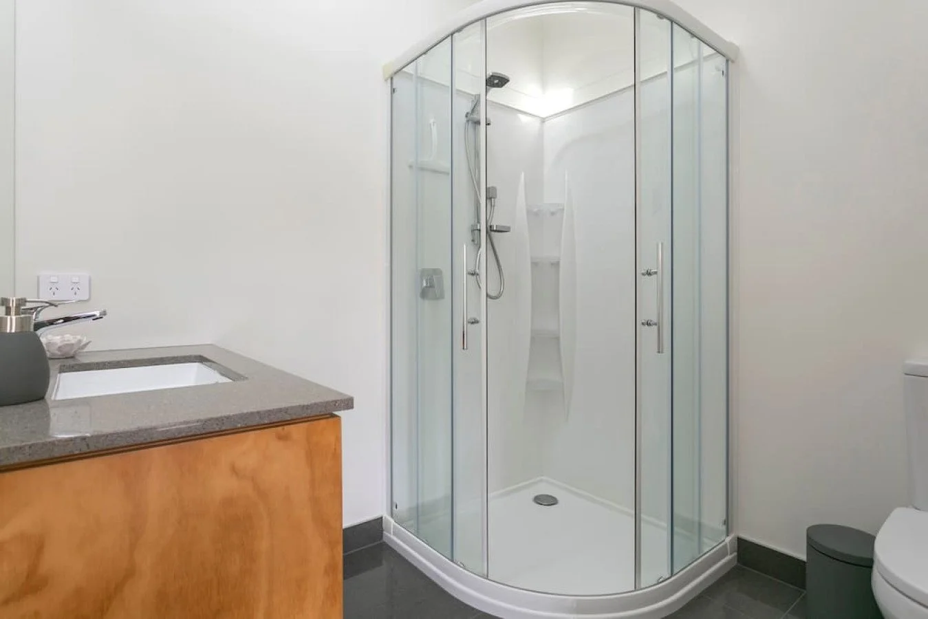 A modern bathroom with a corner glass shower enclosure, a white toilet, a wooden vanity with a gray countertop, a sink, a soap dispenser, and a double electrical outlet on the wall.