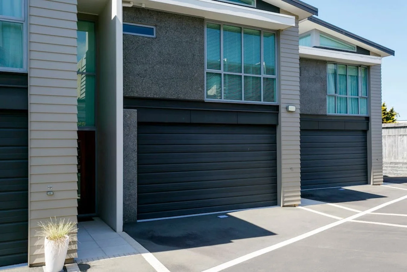 Modern apartment building with multiple black garage doors, large windows with blinds, and a small potted plant near the entrance, sunny day.
