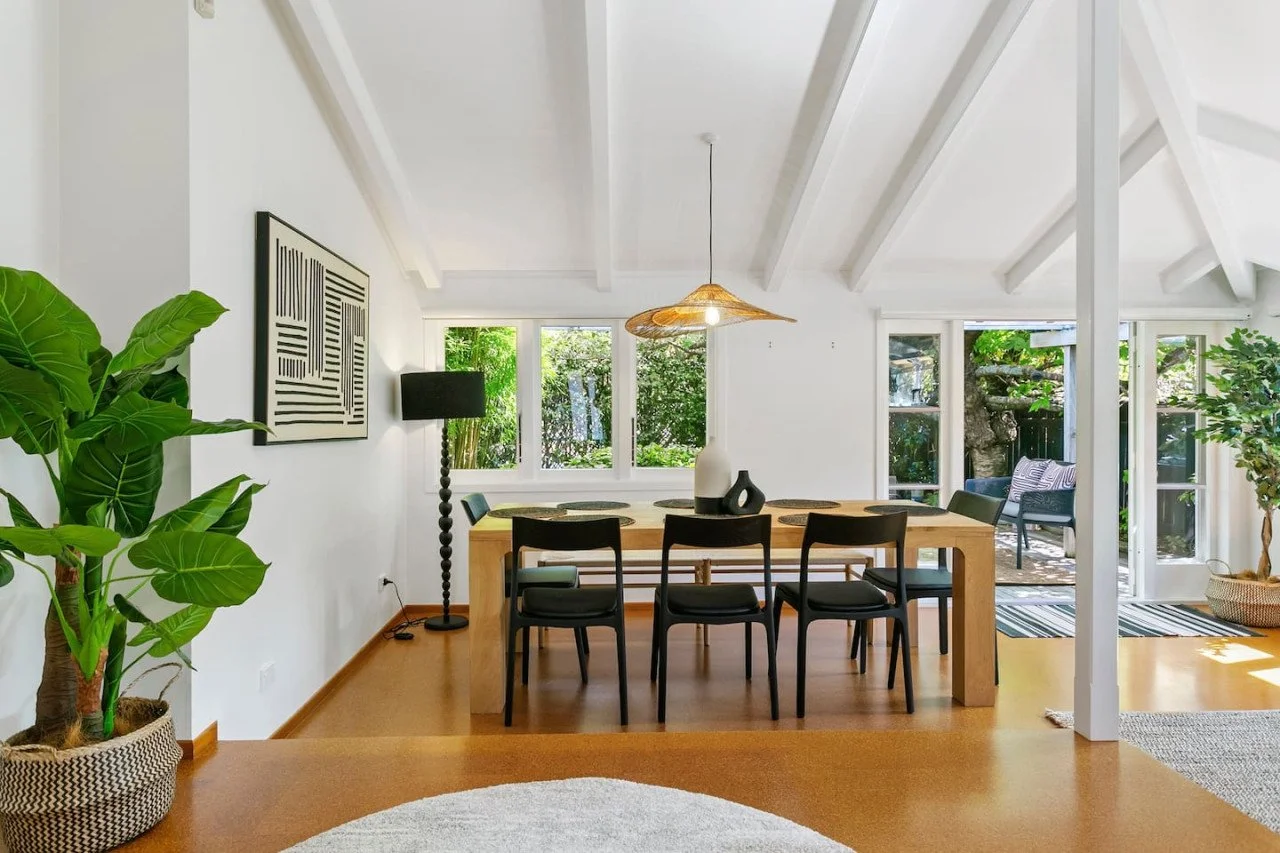 Modern dining room with wooden table, six black chairs, large black floor lamp, white walls, artwork, hanging light fixture, and large windows overlooking a lush green garden with outdoor seating.