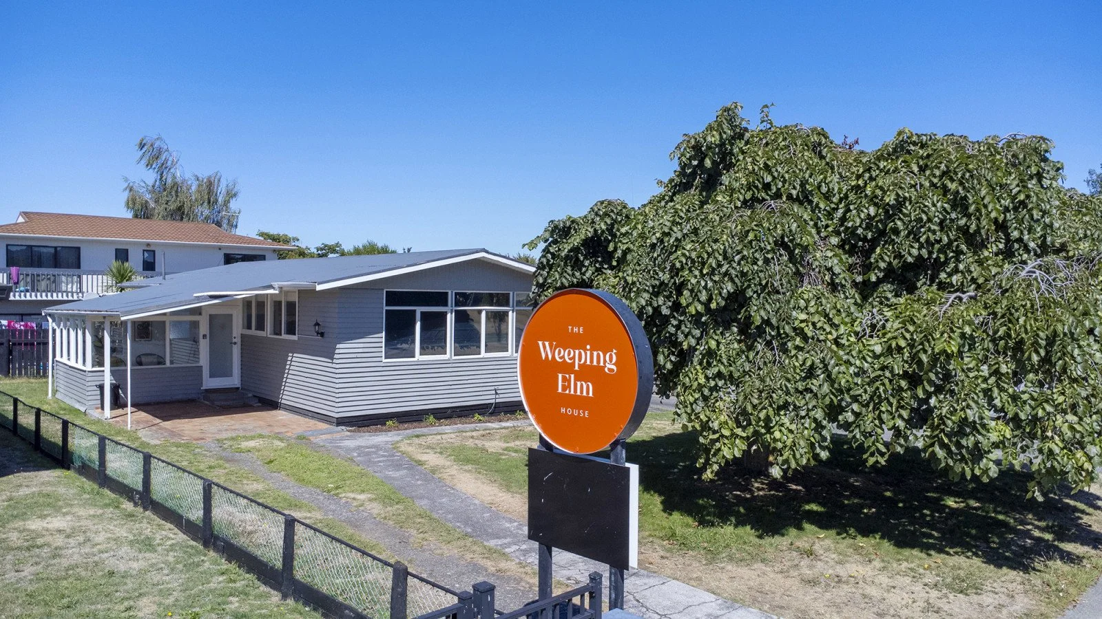 A small house with siding, large front windows, and a covered porch, situated next to a large weeping elm tree, with a sign in the front yard reading 'The Weeping Elm House' under a clear blue sky.