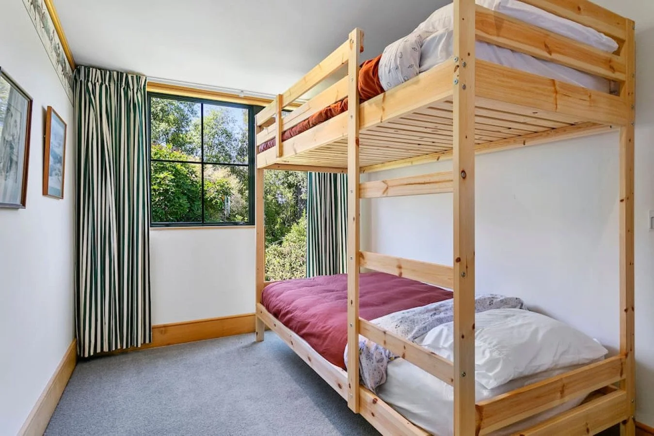 A bedroom with a wooden bunk bed by a window showing green trees outside, with white walls, striped curtains, and framed pictures on the wall.
