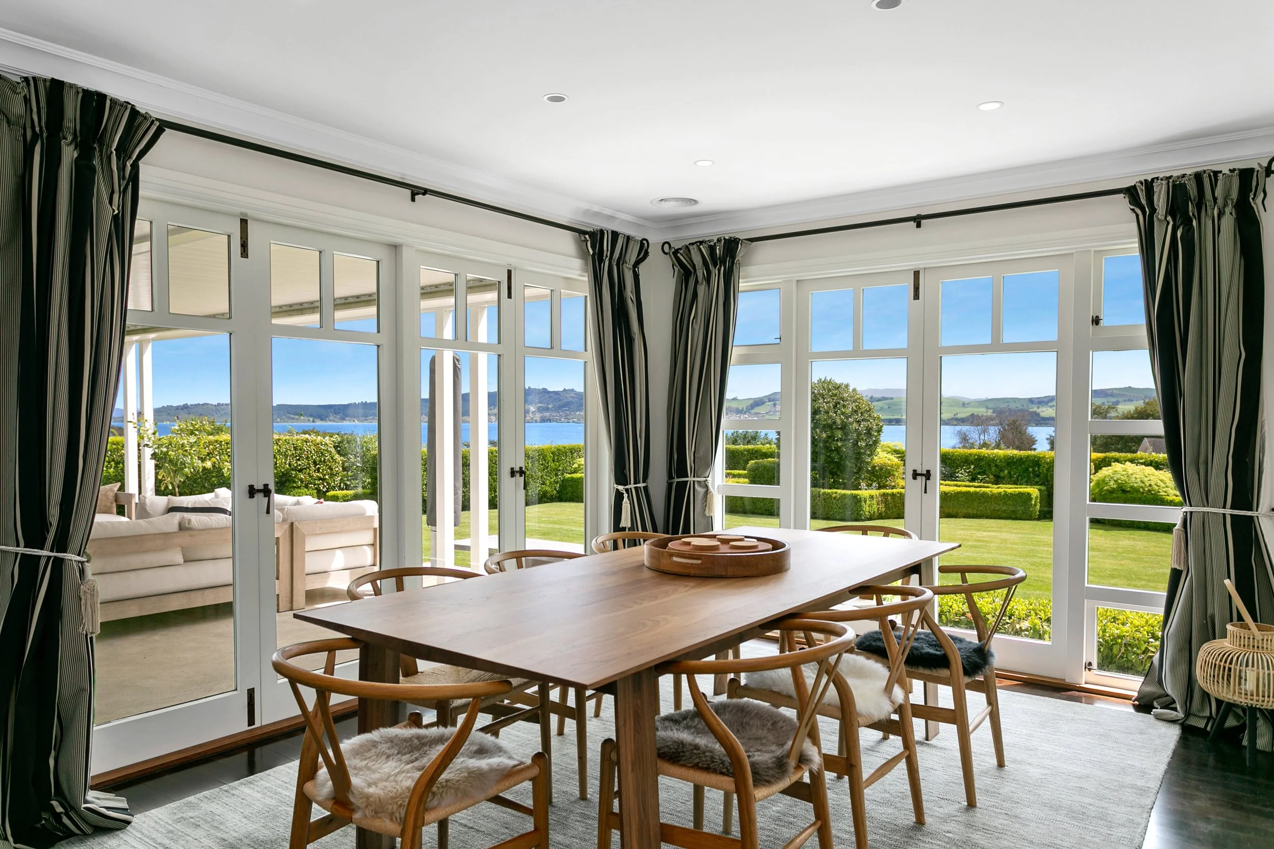 Dining room with wooden table and eight chairs, large windows showing a garden, lake, and hills outside, with curtains pulled back.