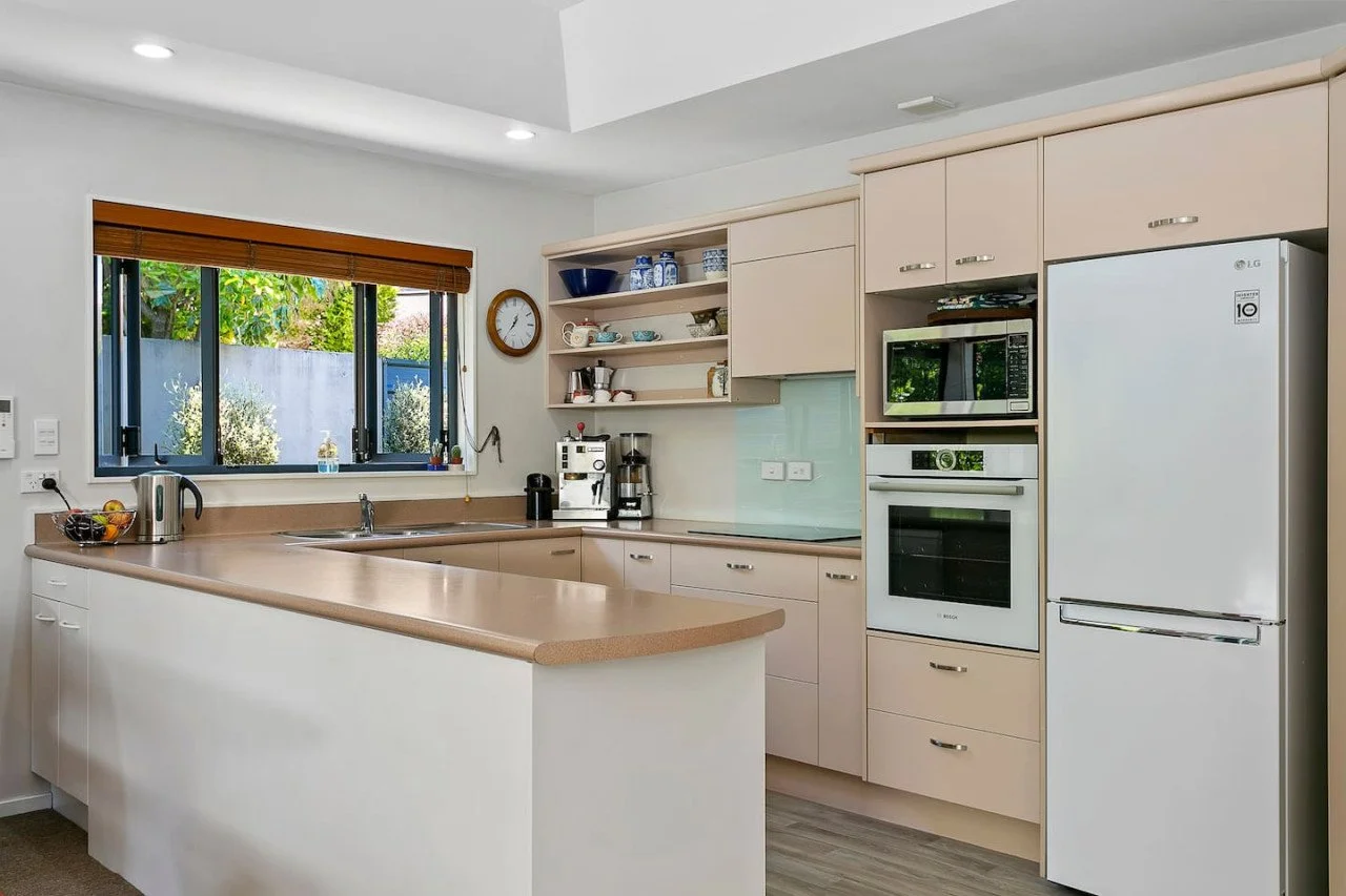 Modern kitchen with beige cabinets, white appliances including a refrigerator, microwave, and oven, a counter with a sink under a window with wooden blinds, open shelves with dishes, and various small appliances on the counter.