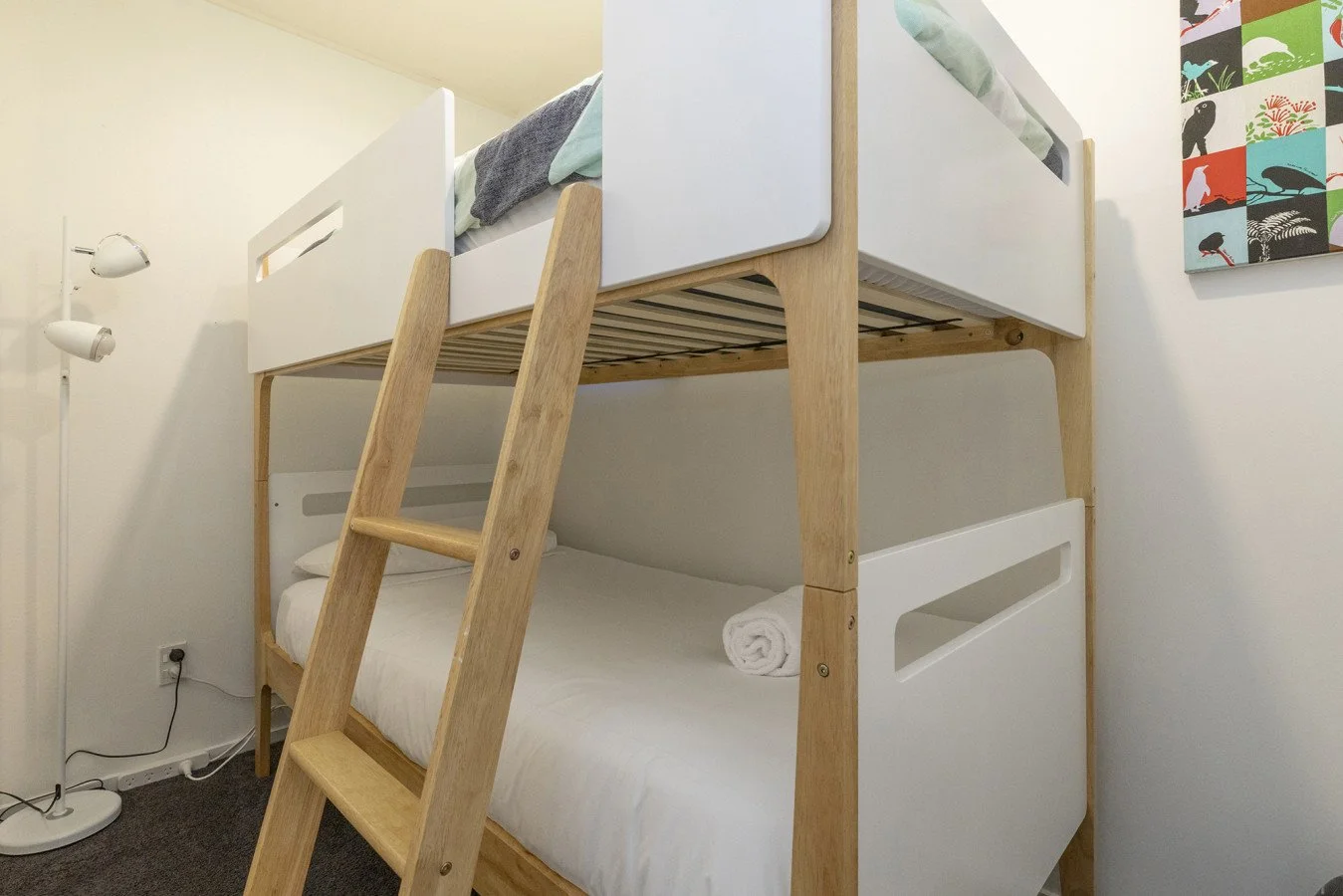 Bunk bed with a wooden frame and white panels, including a lower twin mattress with white linens and a rolled towel, and an upper bed partially visible, located in a room with a white wall, colorful wall art, and a white floor lamp.