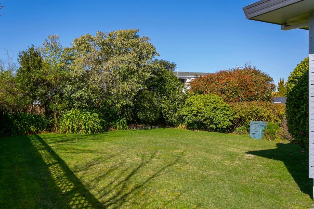 A backyard with well-maintained green grass, surrounded by various bushes and trees under a clear blue sky, with part of a house on the right side of the image.