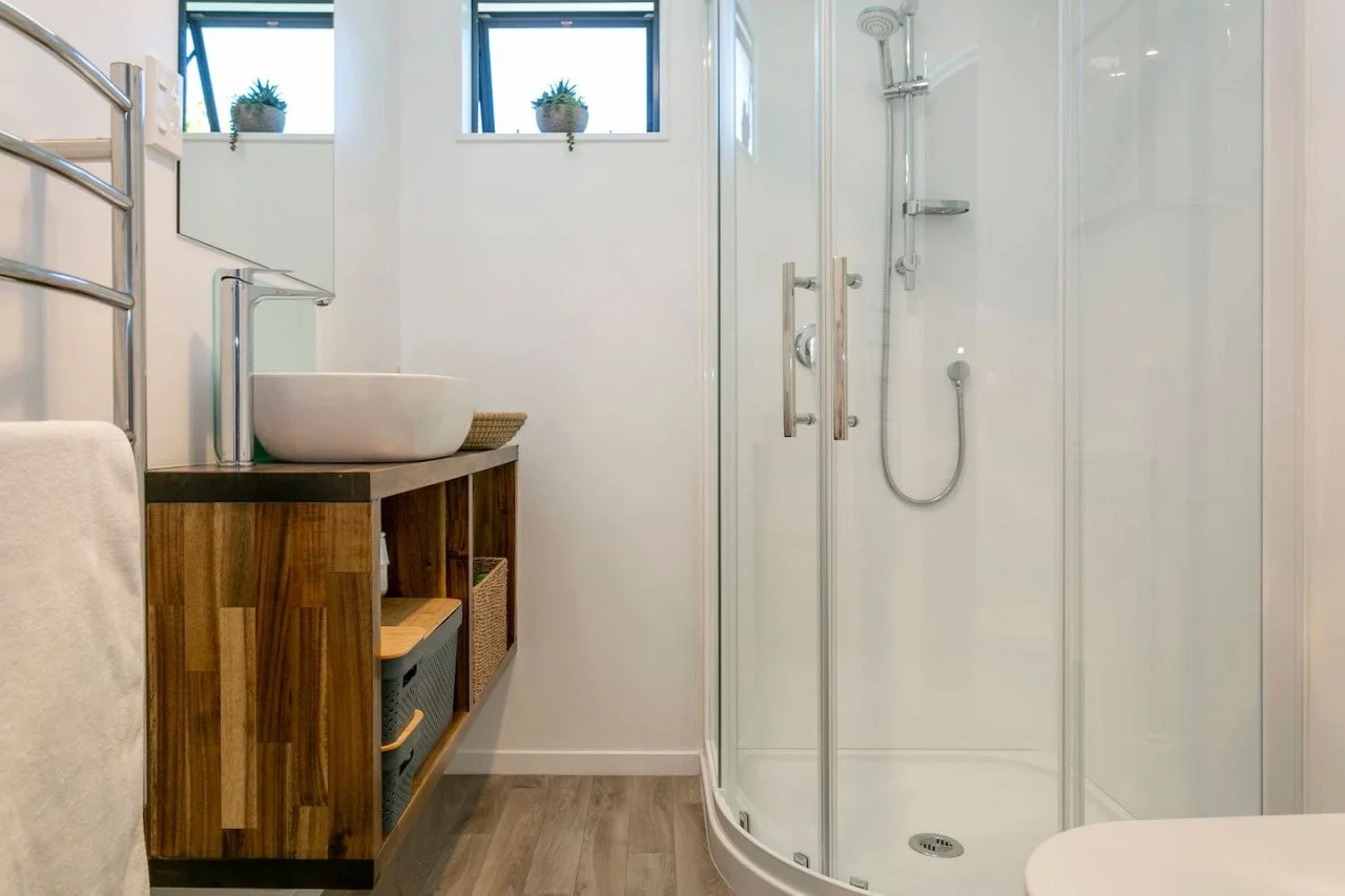 A modern bathroom with a glass shower enclosure, a white vessel sink on a wooden vanity, a towel with a towel rack, and small potted plants on a high window sill.