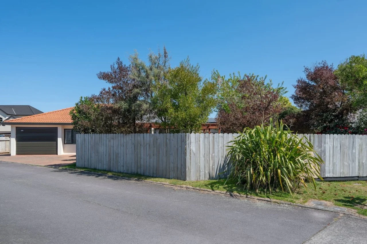 Residential street view showing a house with a garage, a wooden fence, and trees with varied foliage under a clear blue sky.