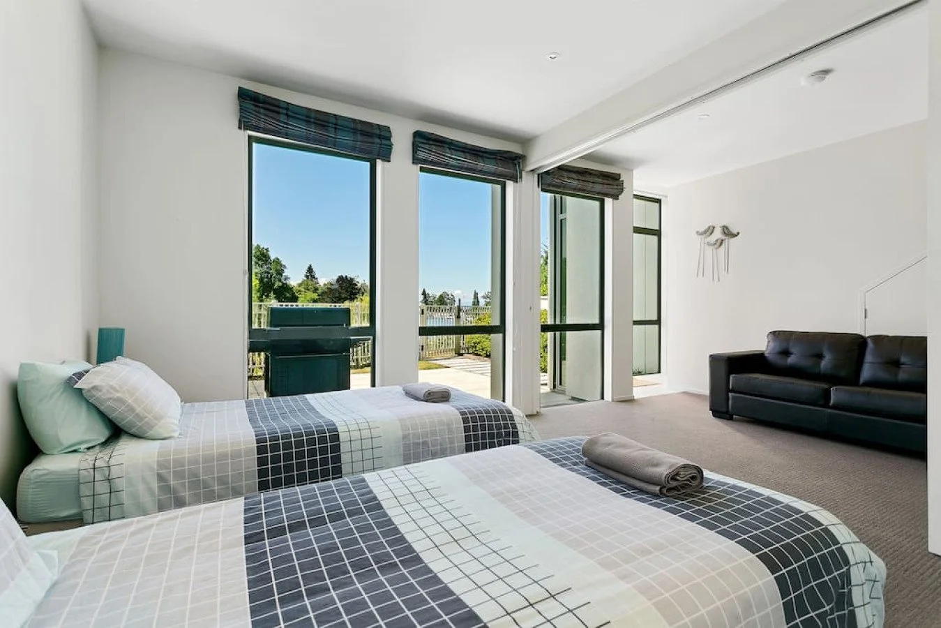 A bright hotel room with two beds, a black sofa, large windows with mountain and sky view, and a small outside patio.
