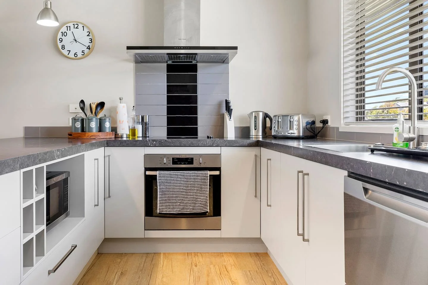 Modern kitchen with white cabinets, black countertop, stainless steel oven, microwave, toaster, kettle, and utensils on the counter. There is a clock on the wall, a window with blinds, and a dish soap near the sink.
