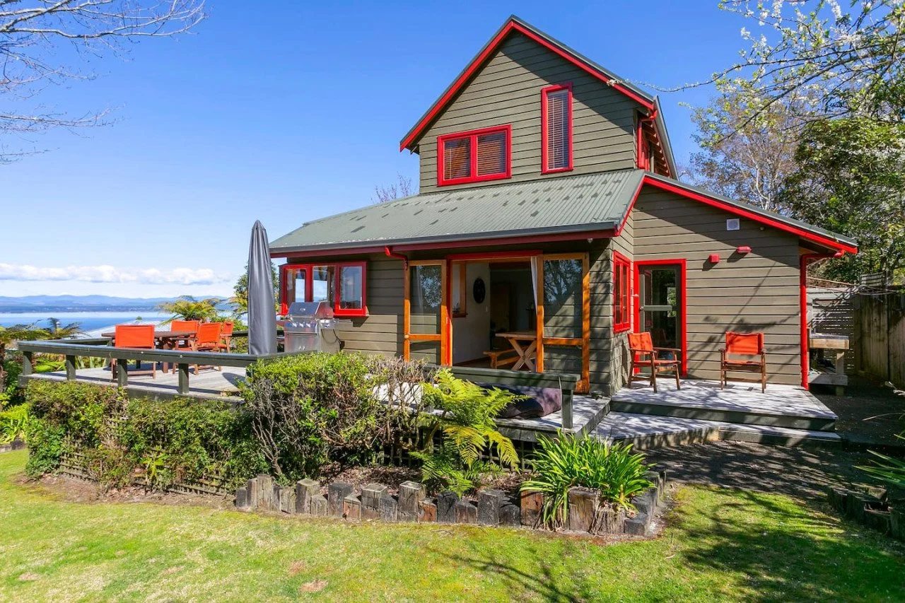 A two-story house with green siding, red window frames, and a green metal roof, surrounded by a well-maintained lawn with a garden, tables, chairs, and a grill on a deck, overlooking a body of water and distant mountains under a blue sky.