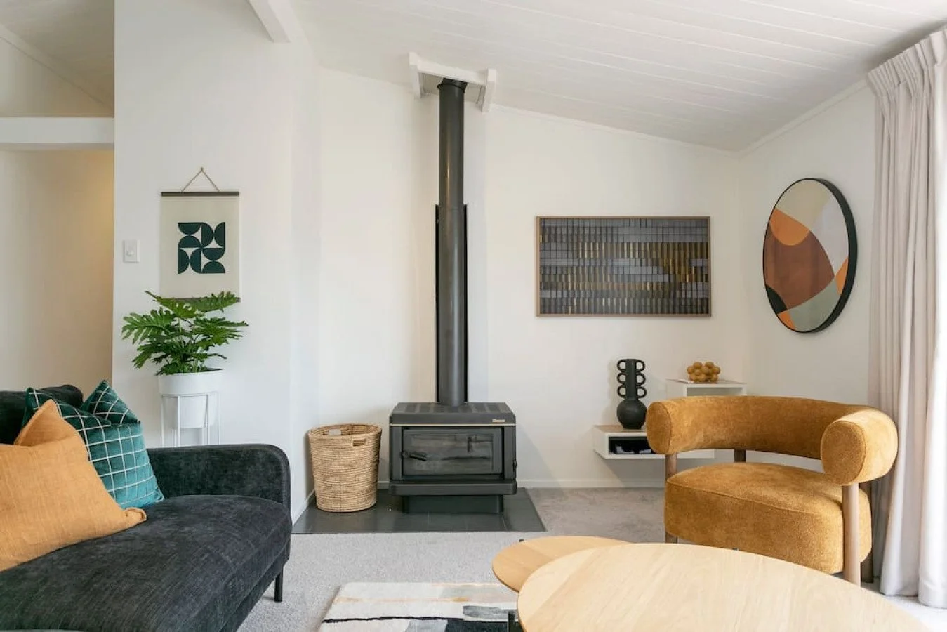 Living room with a wood stove, a black velvet sofa with orange and green pillows, a brass-colored chair, a side table with decorative vases, and wall art, window with white curtains, and a potted plant.