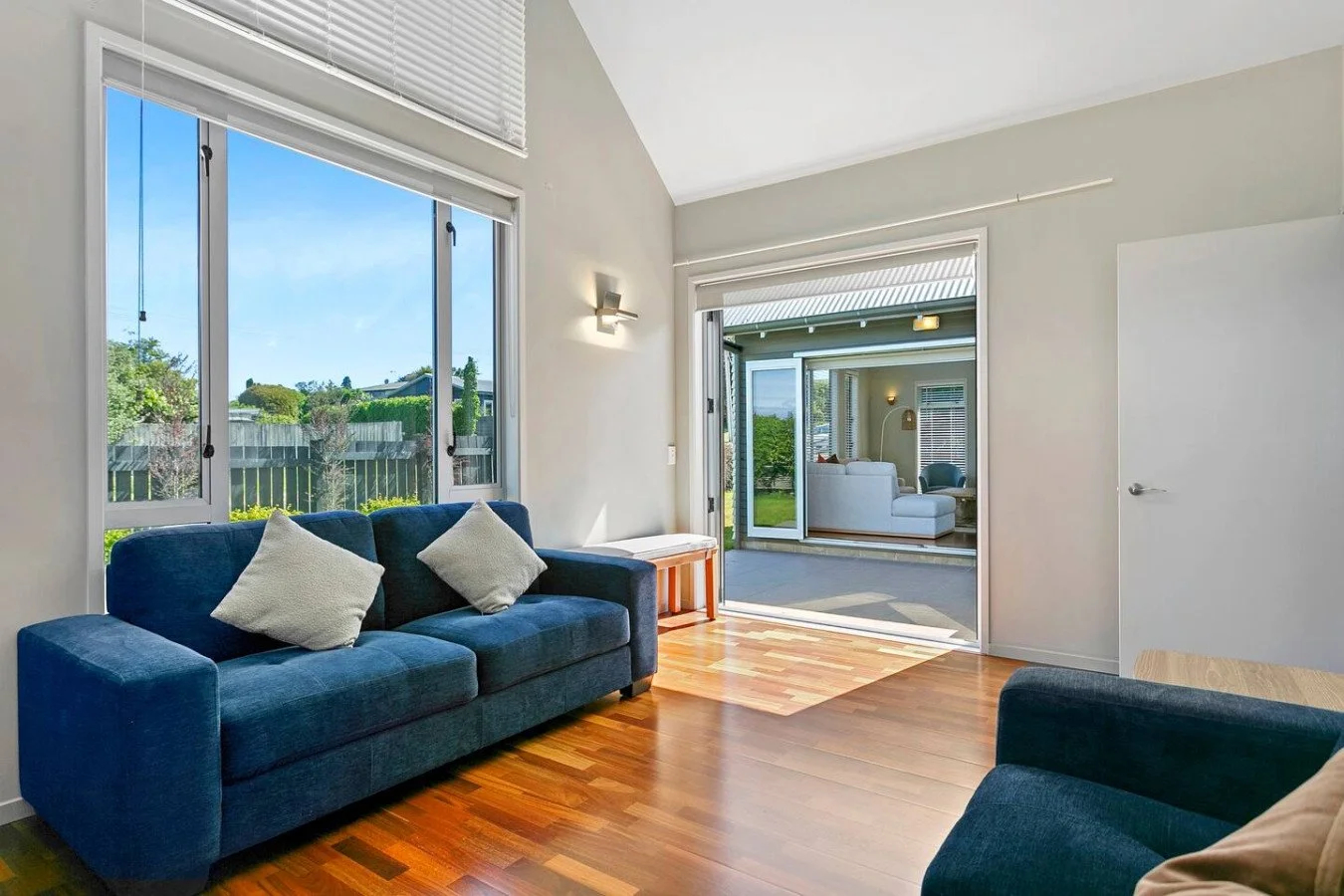 Living room with blue sofa, white pillows, hardwood floors, large windows, and a sliding glass door leading to a patio with outdoor seating and green yard.