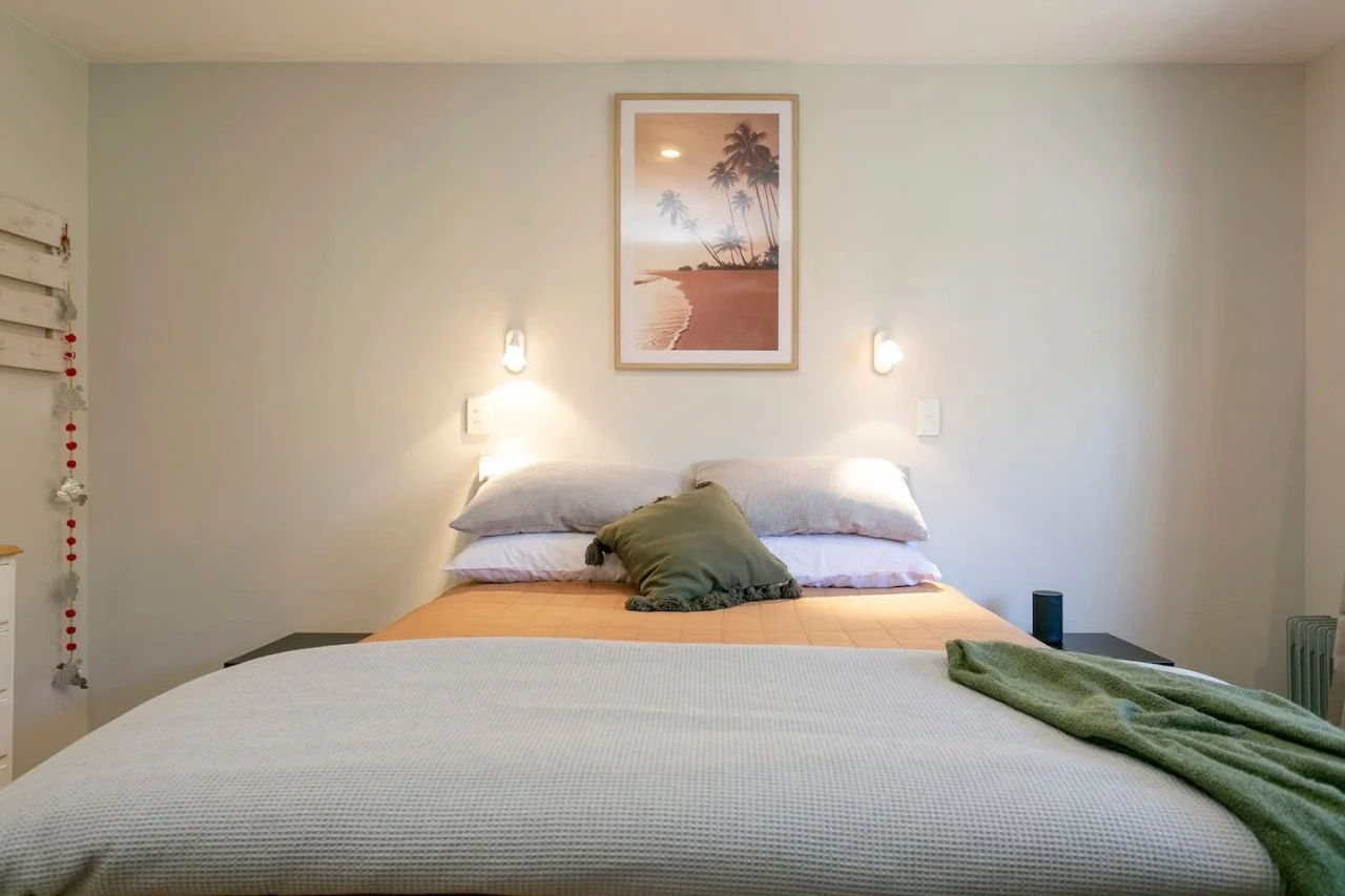 A neatly made bed with white, gray, and green pillows, a green blanket, and a tan bedspread, with a framed beach scene picture and wall-mounted lights above the headboard in a bedroom.