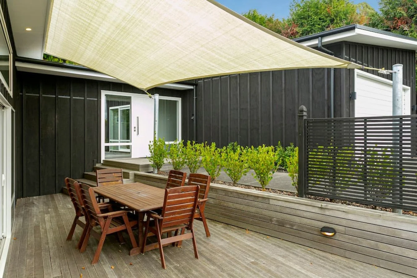 Outdoor patio with wooden table and chairs, a beige sunshade, greenery, black house siding, and a sliding glass door.