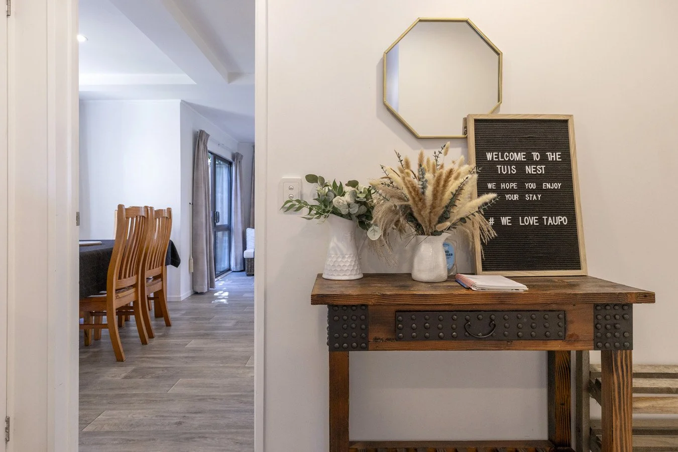 Entryway table with welcome sign, vases of greenery and pampas grass, and decorative mirror in a modern home, with a dining area visible in the background.
