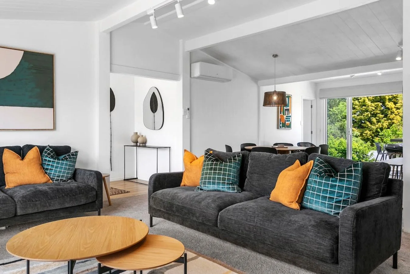 Modern living room with dark gray sofas, colorful pillows, wooden tables, artwork on white walls, large window showing green trees.