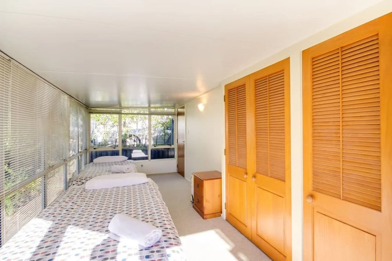 A small room with two beds covered in polka dot bedspreads, a small wooden nightstand, large windows with blinds, wooden closet doors, and a white ceiling.