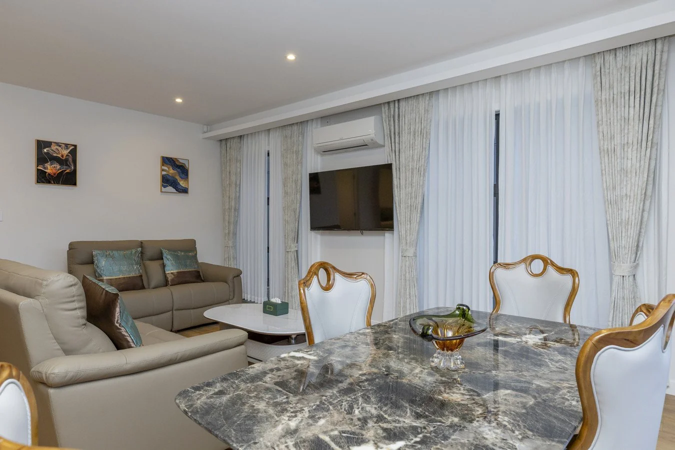 Living room with beige sofas, a marble dining table, white chairs with wooden accents, a wall-mounted TV, and large windows with white curtains.