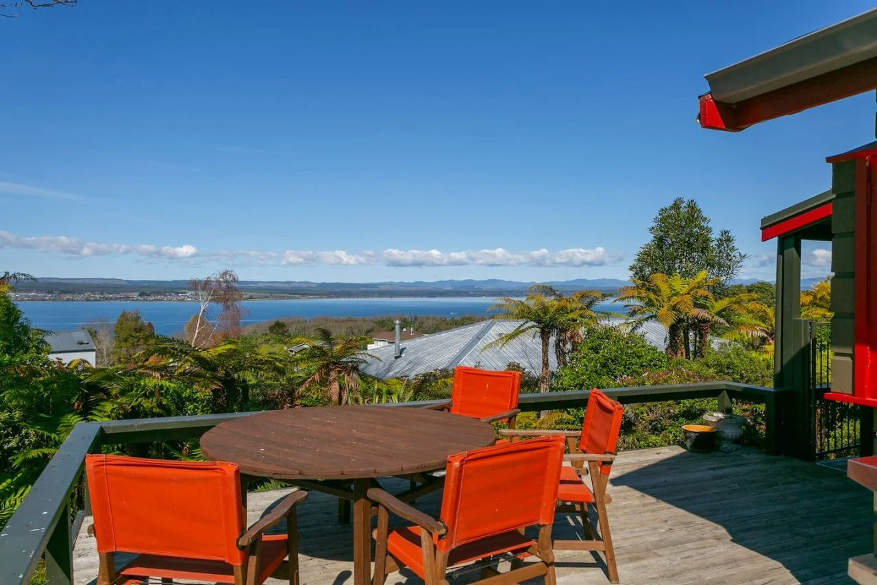 Outdoor patio with a wooden table and four orange chairs, overlooking lush greenery with trees and a lake in the background under a clear blue sky.