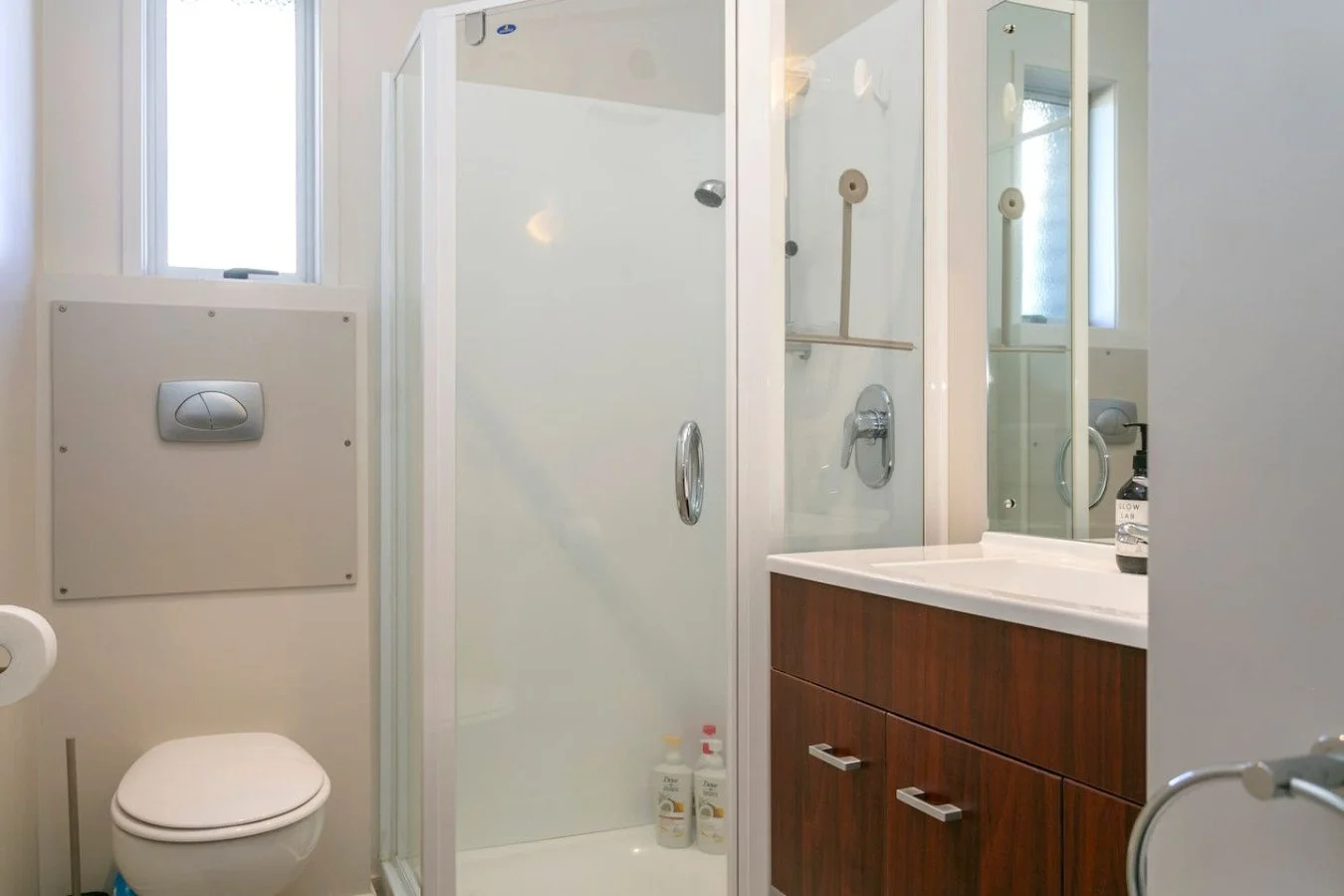 A modern bathroom featuring a white toilet, glass shower with a sliding door, and a wooden vanity with a sink and mirror. There are cleaning supplies inside the shower, and a small window lets in natural light.