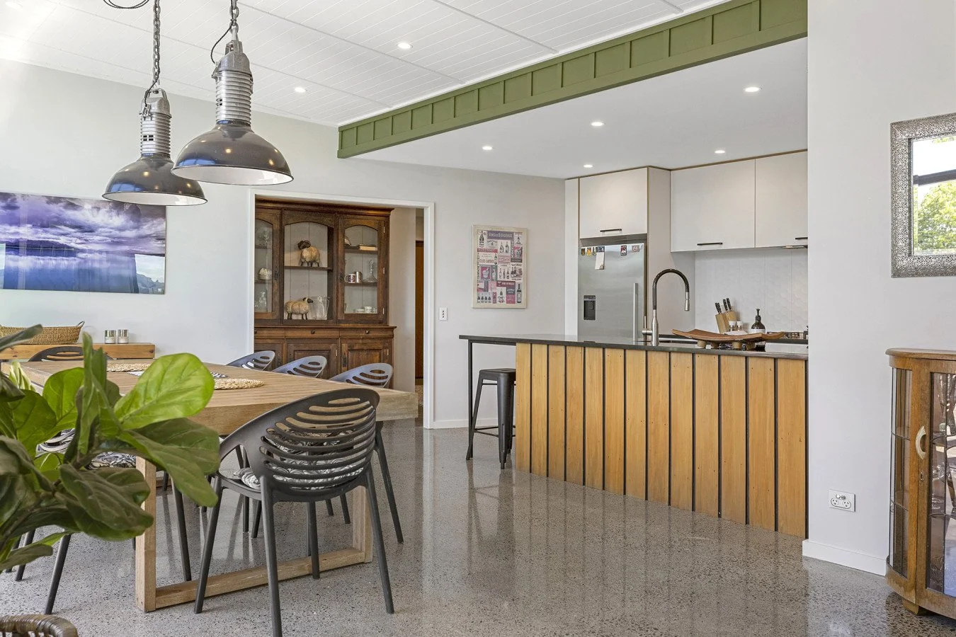 Open-concept kitchen and dining area with modern and rustic decor, including a wooden dining table, black chairs, pendant lights, a kitchen island with wood paneling, and a refrigerator.