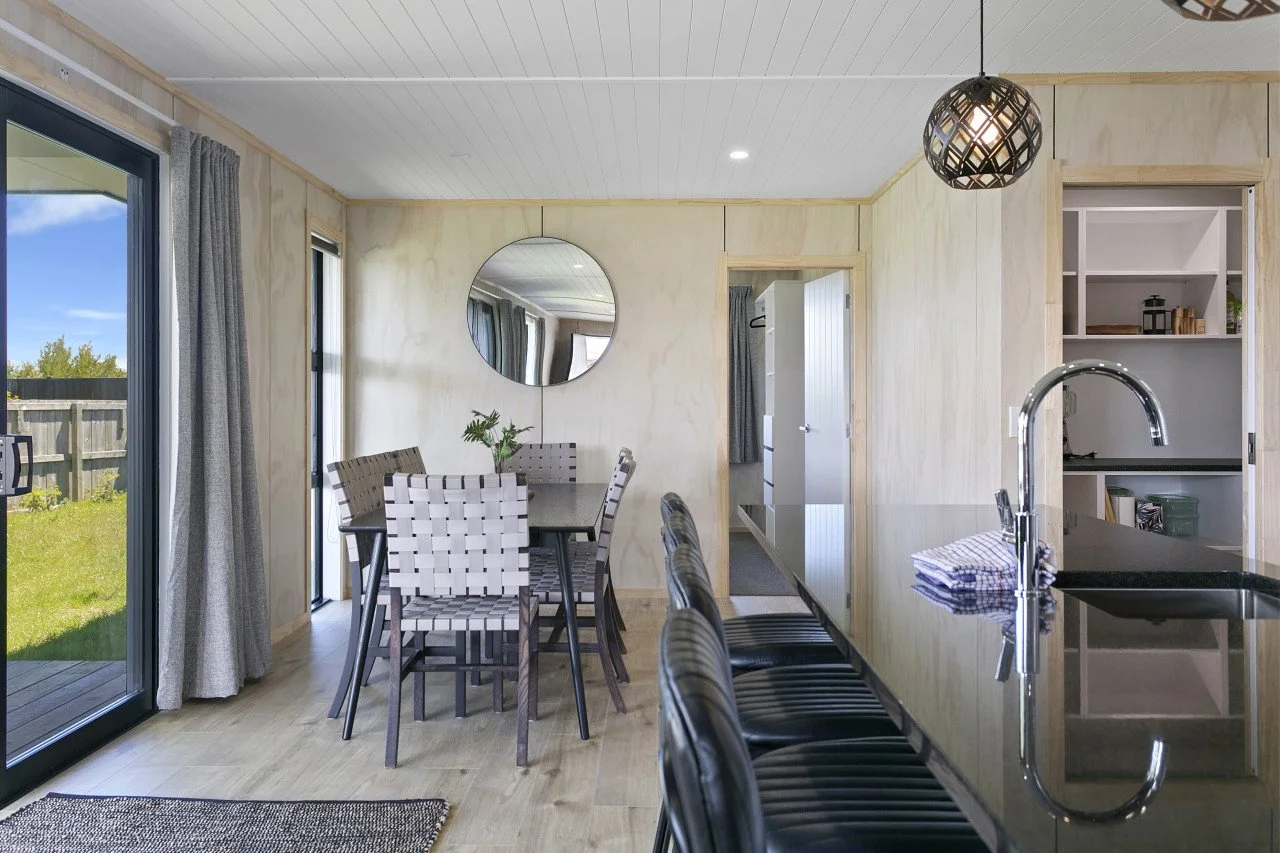 A modern kitchen and dining area with a sliding glass door leading outside, a black granite countertop, and a dining table with six woven style chairs. A round mirror hangs on the wall, and there is a black pendant light fixture. The space has light-