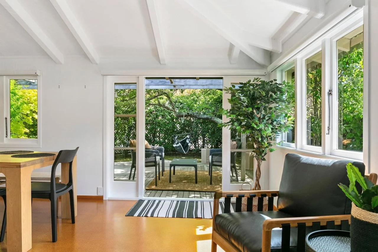 Living room with glass sliding door opening to outdoor patio with chairs, trees, and greenery.
