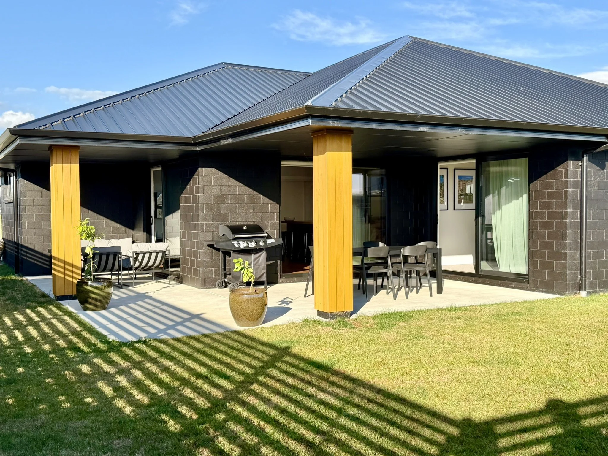 Modern house with black brick exterior, metal roof, and outdoor patio with seating, potted plants, and a barbecue grill. Green lawn and shadow pattern from fencing under a blue sky.