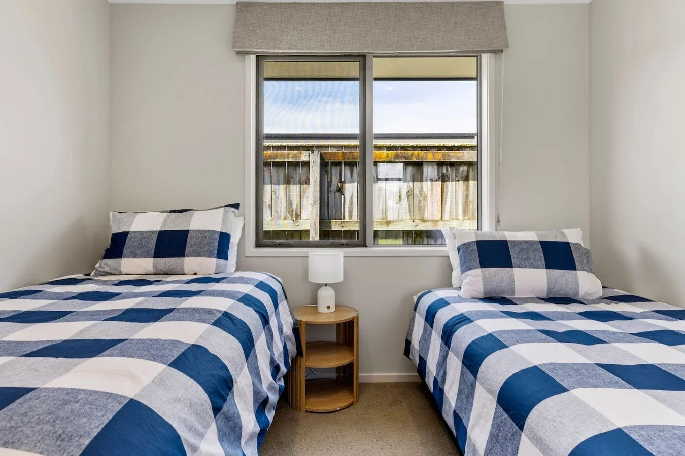 A bedroom with two twin beds with blue and white checkered bedding, a small wooden nightstand between them, and a window with a view of a wooden fence outside.