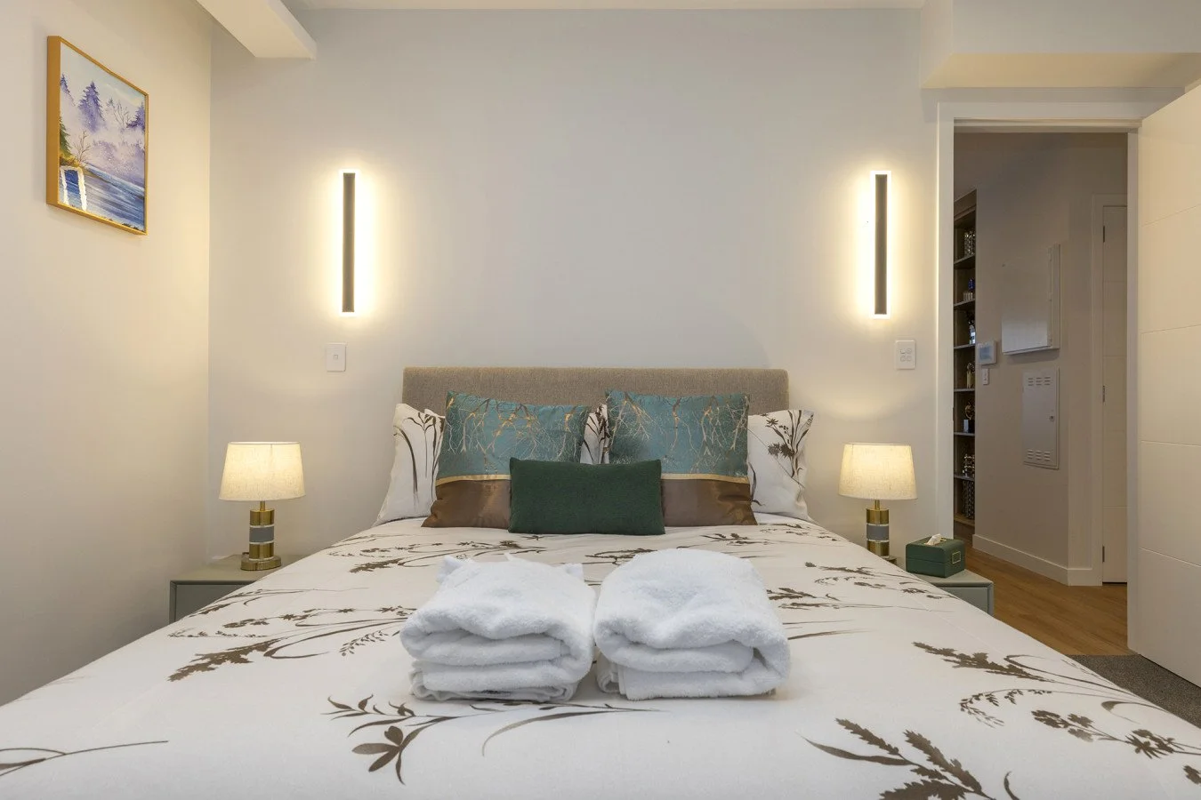 A neatly made bed with rolled white towels on top, decorative pillows in shades of green and cream, bedside lamps, and modern wall lighting in a contemporary bedroom.