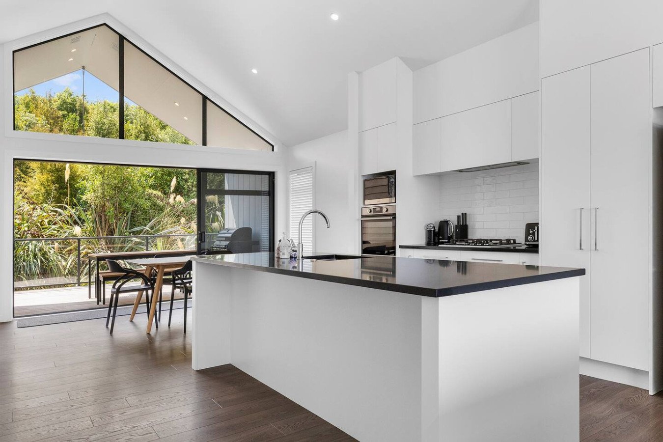 Modern kitchen with white cabinets, black countertop island, stainless steel appliances, and large windows with a view of greenery, dining table, and outdoor balcony.