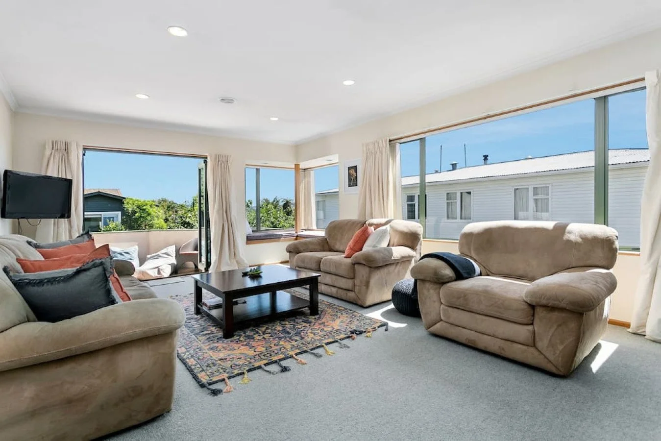 Bright living room with beige sofas, a dark wooden coffee table, a colorful patterned rug, and large windows with white curtains showing a blue sky and neighboring houses.