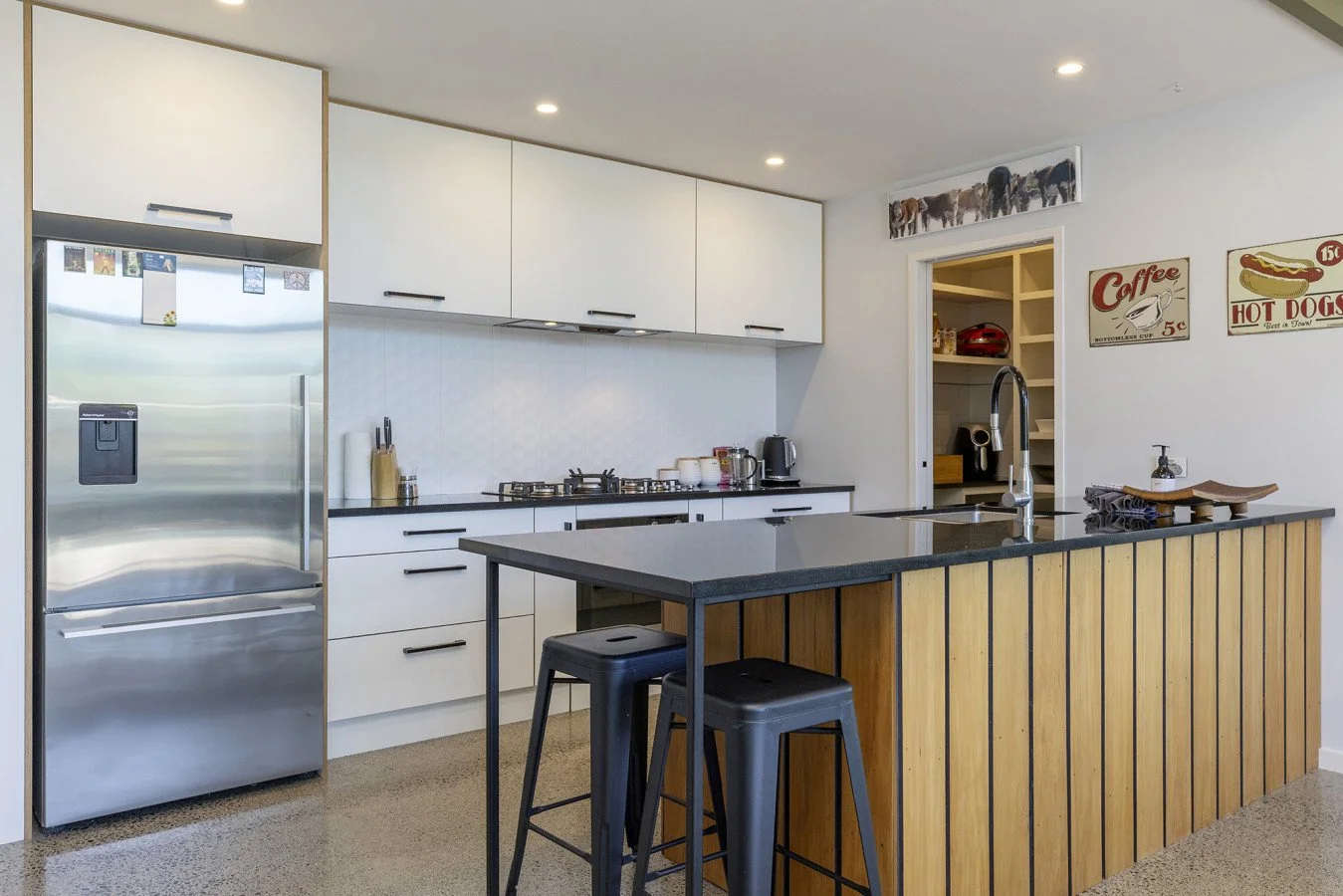 Modern kitchen with white cabinets, black countertops, stainless steel refrigerator, island with black countertop and wooden panel, two black bar stools, kitchen appliances, and decorative signs on the wall.