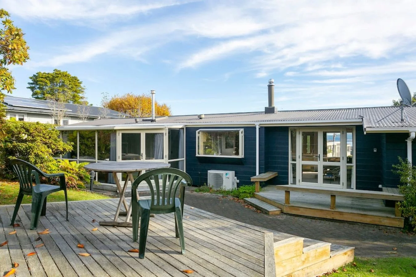 A backyard with a wooden deck, plastic chairs, a table, green grass, bushes, and a blue house with a screened porch and glass doors, under a partly cloudy sky.