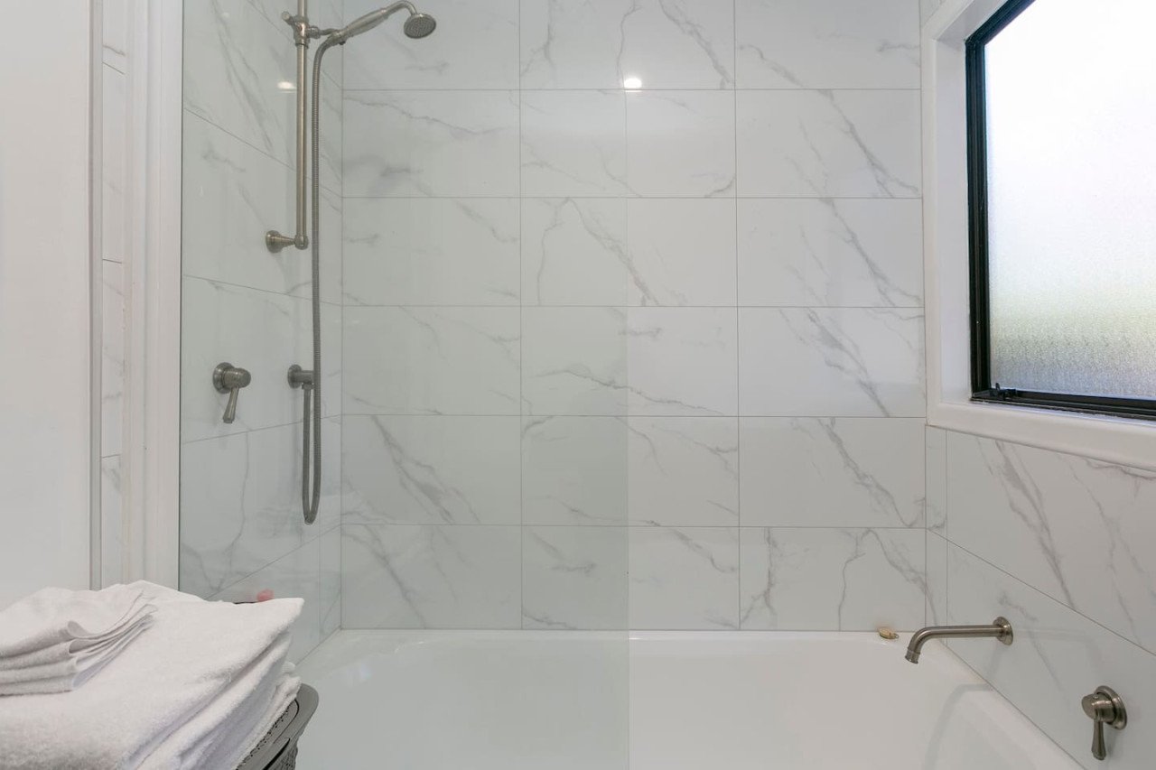 Bathroom shower with marble wall tiles, a frosted window, a showerhead, a handheld shower, and a bathtub with a wall-mounted faucet, with folded towels on a small table.