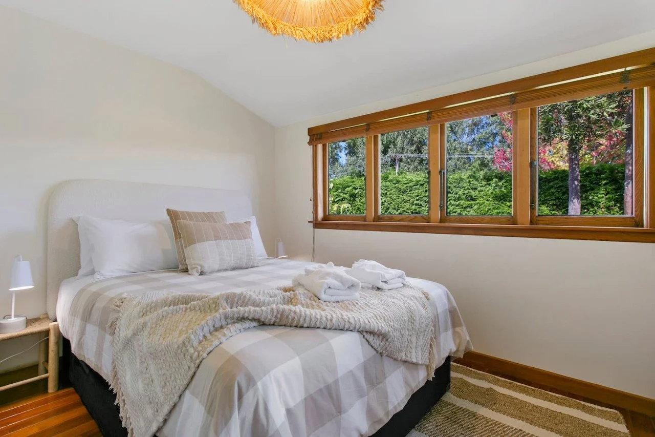 Cozy bedroom with white walls, a bed with beige striped bedding, a beige pillow, and folded towels, large windows with wooden blinds showing outdoor greenery and trees.