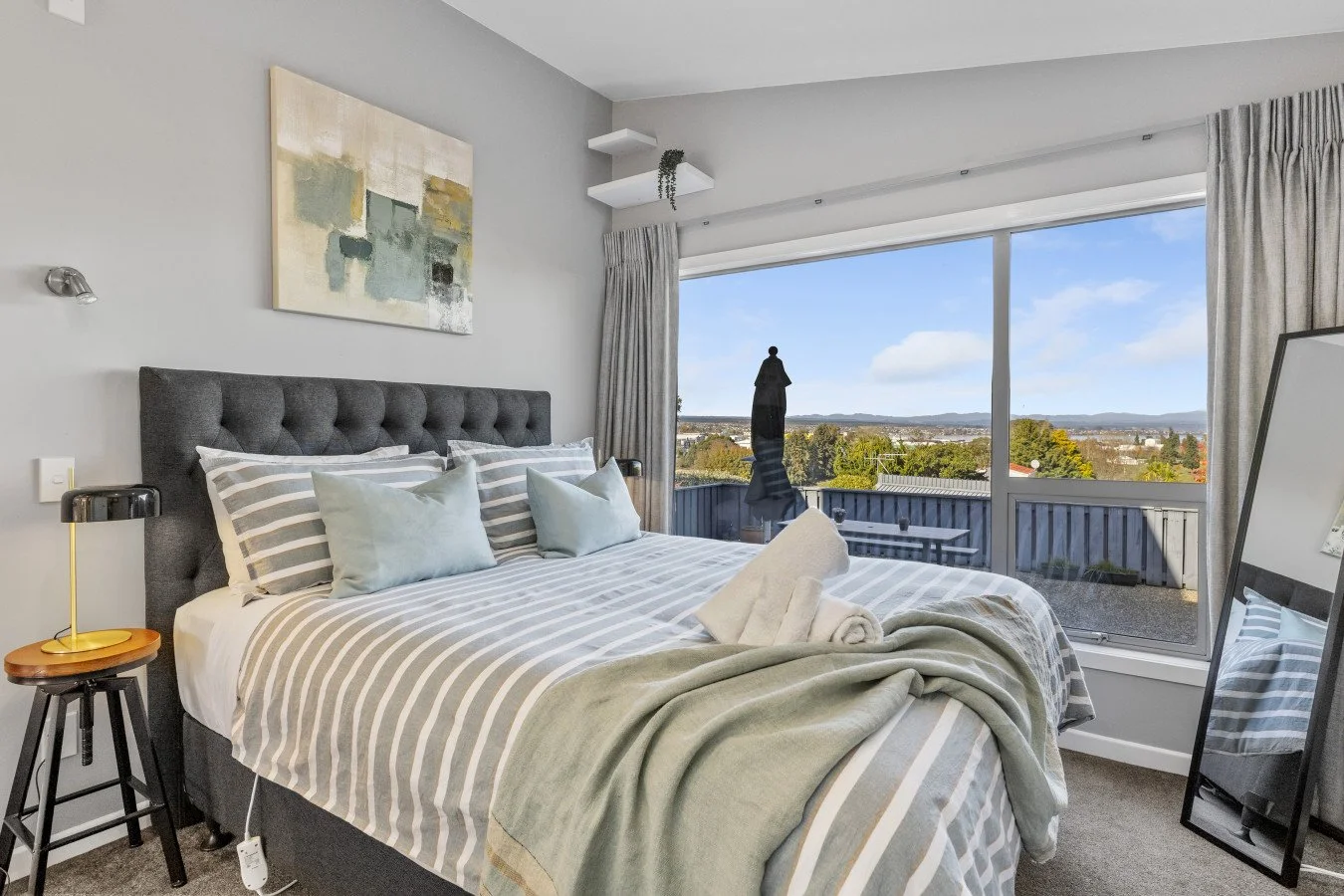 A modern bedroom with a large bed, gray tufted headboard, striped bedding, pillows, and a green throw blanket, next to a large window with city and mountain view.