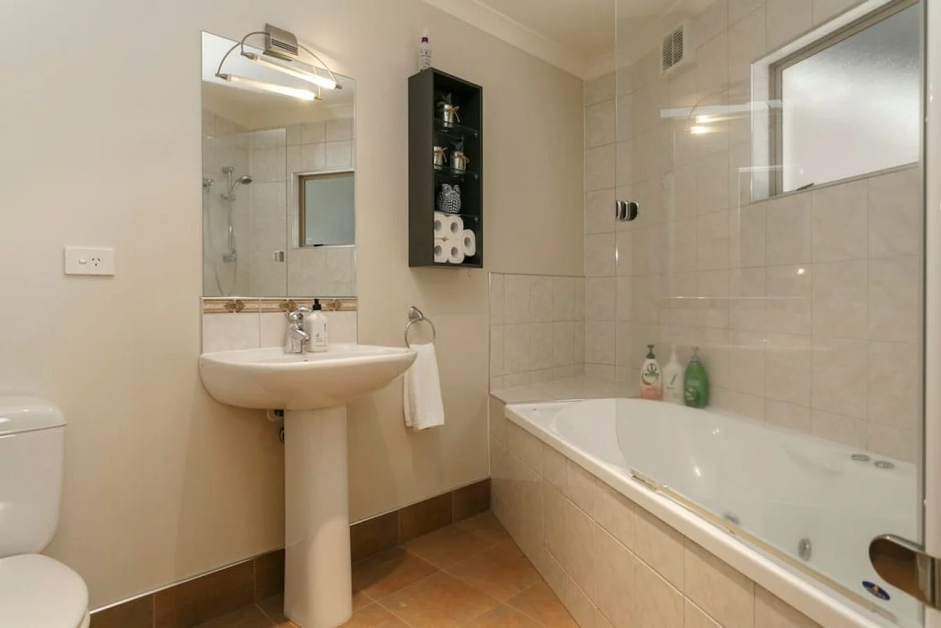 Bathroom with a white pedestal sink, a mirror, a tub with bottled soap, a small wall shelf holding toiletries and toilet paper, a window, and a shower area in the background.