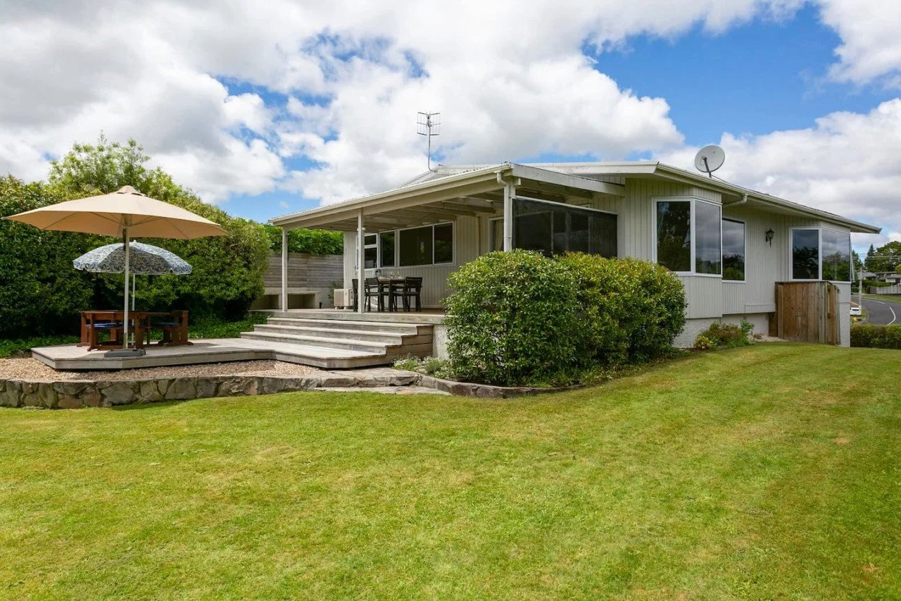 A house with a white exterior and large windows, a front porch with steps, a wooden deck with patio furniture, and a grassy yard with bushes and trees, under a partly cloudy sky.