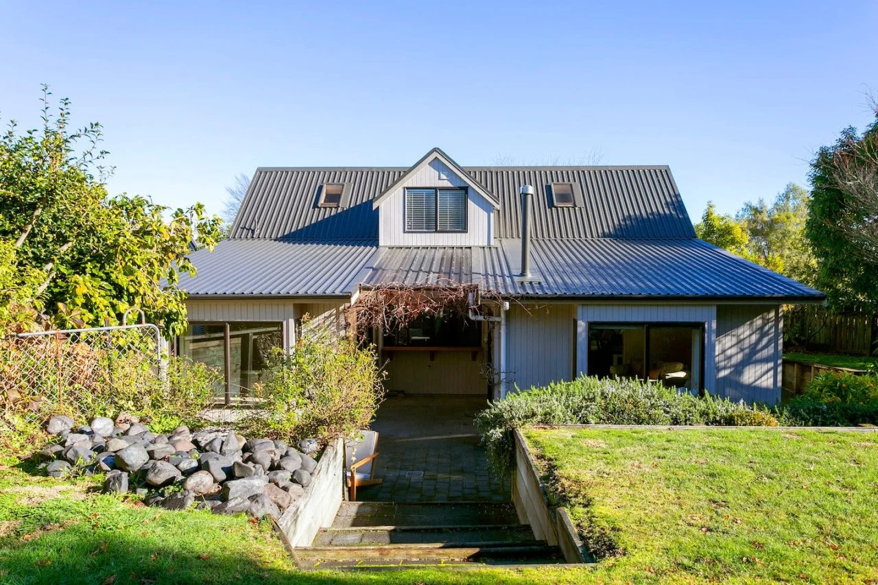 Front view of a house with metal roof, large glass sliding doors, and a small yard with garden beds and a lawn.