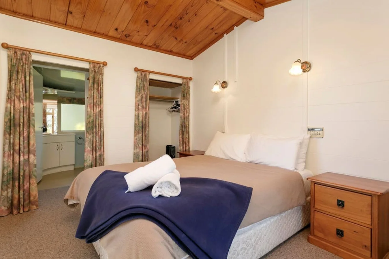 A cozy bedroom with wooden vaulted ceiling, white walls, and a bed with white pillows, a beige blanket, and a navy blue throw at the foot. Two evening wall lamps are above the bed, and a wooden nightstand is on the side. There are two windows with fl