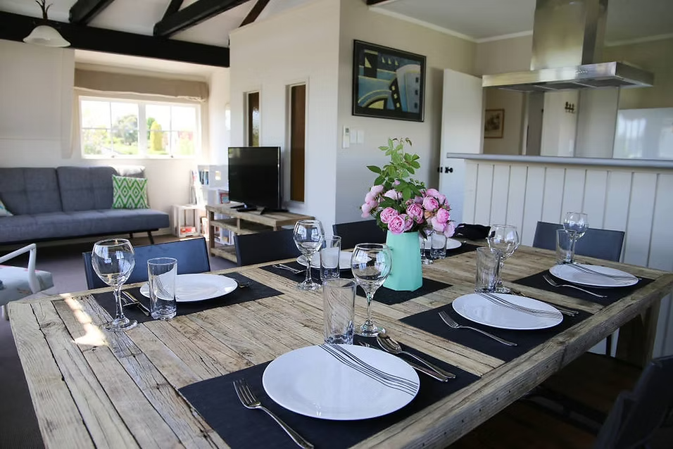 A dining table set with six place settings, including white plates, silverware, and water glasses, with a turquoise vase filled with pink roses as a centerpiece. The room is a bright, open living space with a gray couch, TV, and artwork on the walls.