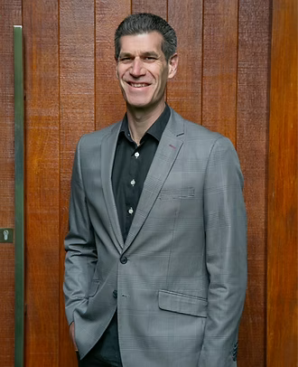 A man in a gray suit smiling, standing against a wooden wall background.