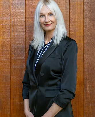 A woman with silver hair wearing a black blazer and striped shirt, standing in front of a wooden wall.