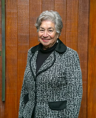 An elderly woman with gray hair, smiling, standing in front of a wooden wall. She is wearing a black turtleneck, a patterned blazer, and earrings.